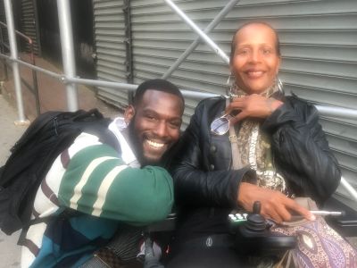 A Black woman in a wheelchair smiling. To her left is a Black man who is also smiling.
