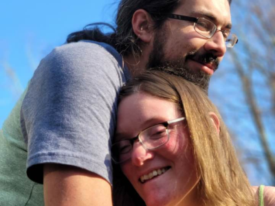 A man and a woman wearing glasses. They are embracing.